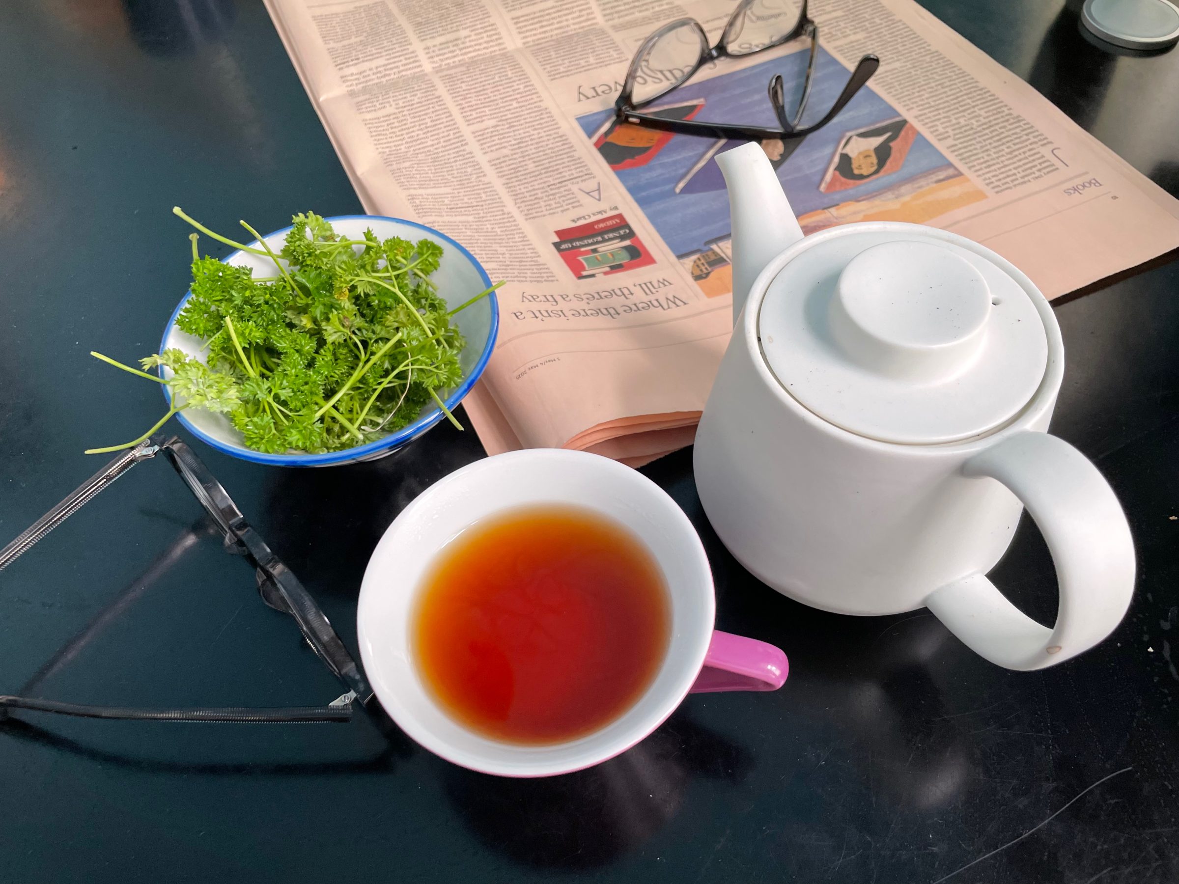 Eine weiße Teekanne und eine Tasse Tee auf einem dunklen Tisch, daneben eine Schale Petersilie, zwei Brillen und eine aufgeschlagene Zeitung.