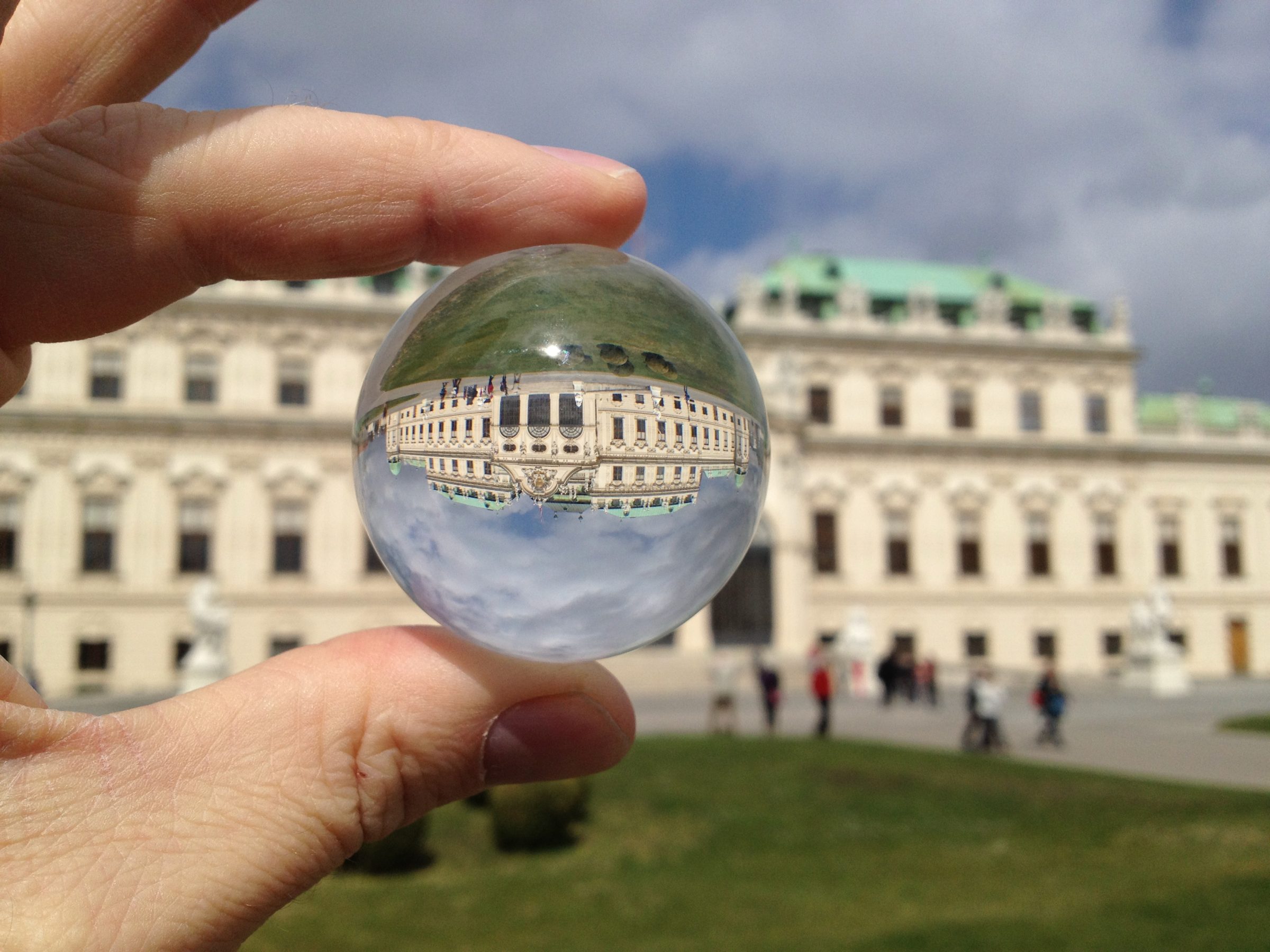 Eine Glaskugel zwischen zwei Fingern; in ihr steht das Obere Belvedere in Wien auf dem Kopf, dahinter das Schloss selbst.